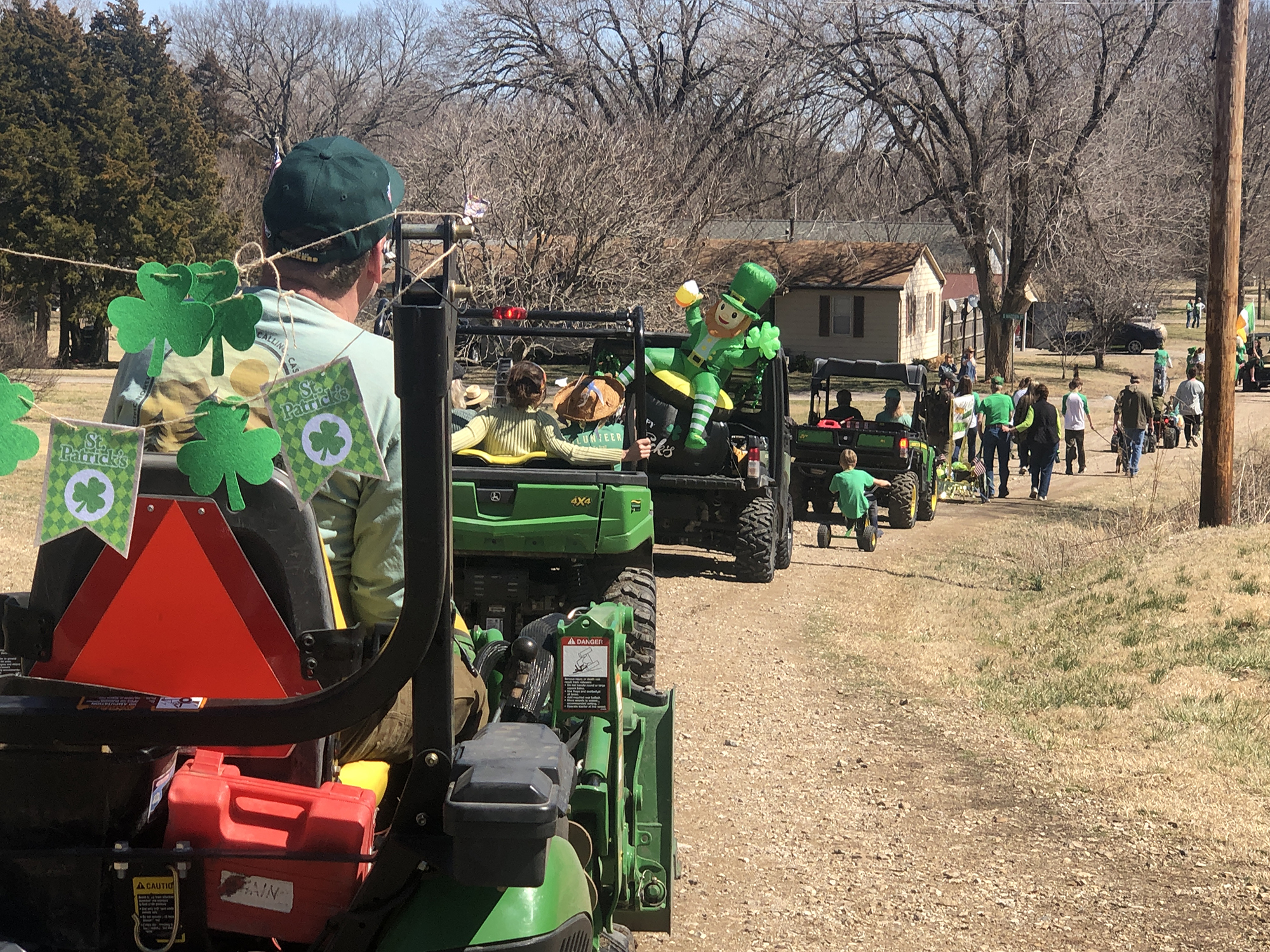 A group people drive assorted green tractors drive down a dirt road. People in the tractors are dressed for St. Patrick's Day Parade in 2023.